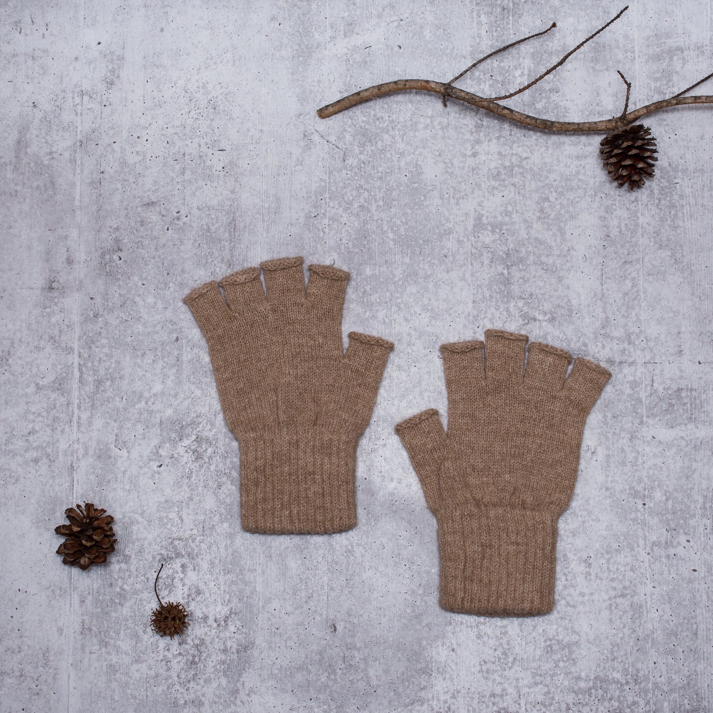 Pair of brown fingerless gloves on a gray surface with pine cones and a branch.