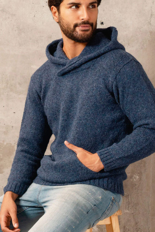 Man wearing a blue knitted sweater sitting on a wooden stool against a concrete wall.